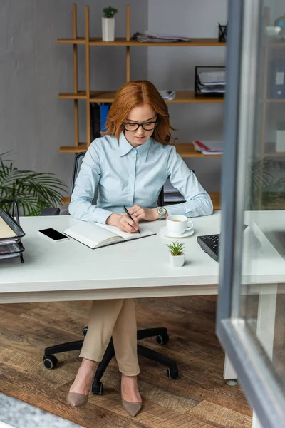 Toute la longueur de l'écriture femme d'affaires dans un cahier, tout en étant assis sur le lieu de travail avec fenêtre floue au premier plan — Photo de stock