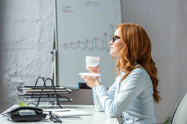 Vue latérale de la femme d'affaires avec soucoupe et tasse de café regardant loin, tout en étant assis sur le lieu de travail sur fond flou — Photo de stock