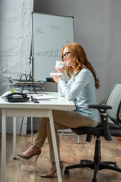 Lunghezza completa della donna d'affari rossa con tazza di caffè e piattino seduto sul posto di lavoro con lavagna a fogli mobili sullo sfondo — Foto stock