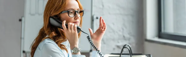 Pelirroja mujer de negocios haciendo gestos mientras habla por teléfono fijo con un rotafolio borroso en el fondo, pancarta - foto de stock