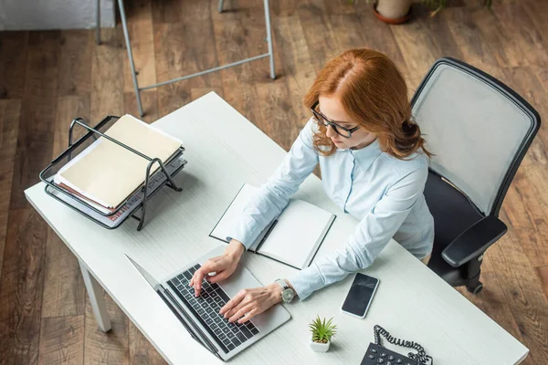 Visão geral da mulher de negócios ruiva digitando no laptop, enquanto está sentado no local de trabalho com dispositivos — Fotografia de Stock