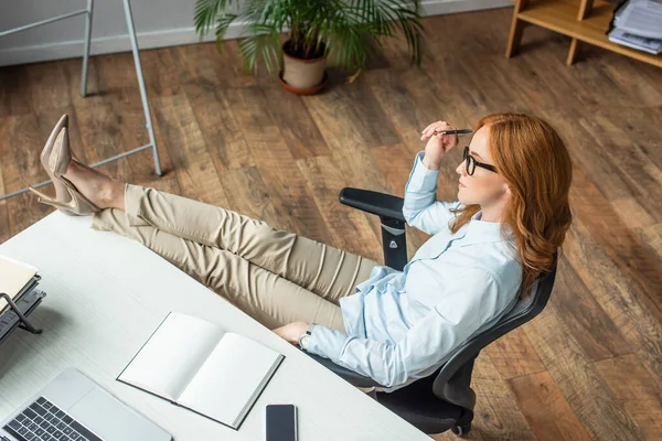 Вид сверху на рыжую деловую женщину со скрещенными ногами, сидящую рядом с рабочим местом с цифровыми устройствами — стоковое фото