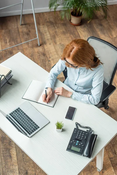 Vue aérienne rousse femme d'affaires écrivant dans un cahier, tout en étant assis sur le lieu de travail avec des appareils — Photo de stock