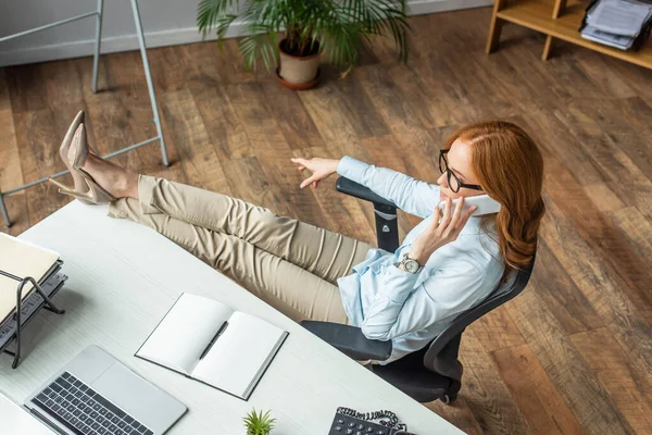 Vista ad alto angolo di donna d'affari rossa con gambe incrociate sul tavolo che parla sul telefono cellulare in ufficio — Foto stock