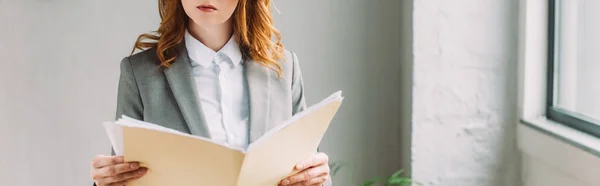 Cropped view of redhead businesswoman holding folder with paper sheets near window, banner — Stock Photo