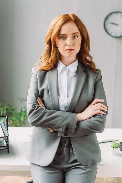Vista frontal de una mujer de negocios seria con brazos cruzados mirando la cámara cerca del lugar de trabajo sobre un fondo borroso - foto de stock