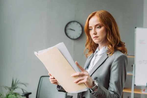 Seriöse Geschäftsfrau blickt auf Ordner mit Dokumenten mit verschwommenem Büro im Hintergrund — Stockfoto