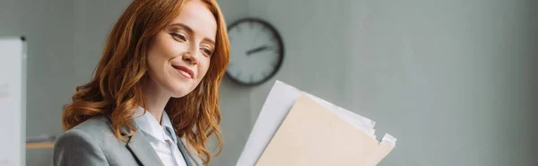 Mujer de negocios positiva mirando a la carpeta con hojas de papel con reloj de pared borrosa en el fondo, bandera - foto de stock