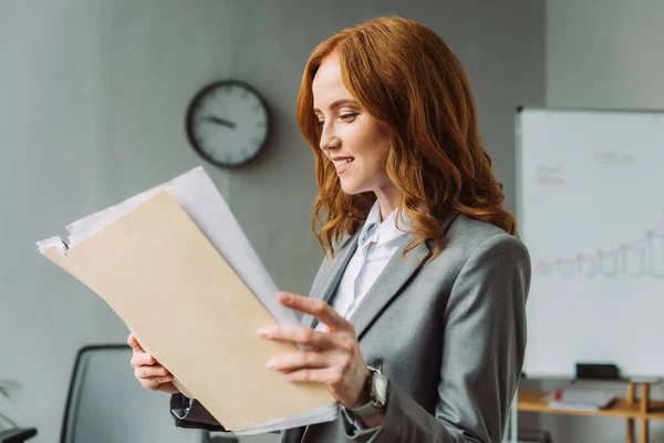 Smiling redhead businesswoman looking at folder with documents with blurred office on background — Stock Photo