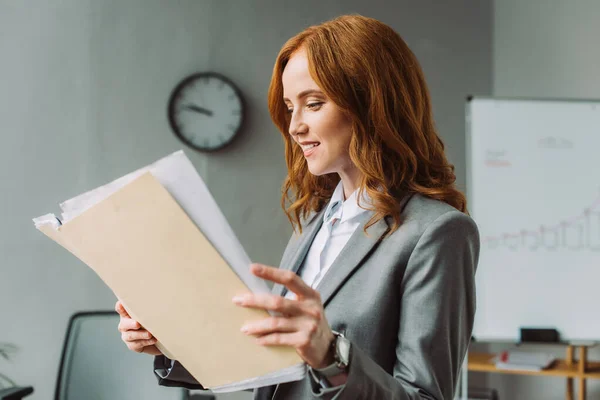 Femme d'affaires rousse souriante regardant le dossier avec des documents avec un bureau flou sur fond — Photo de stock