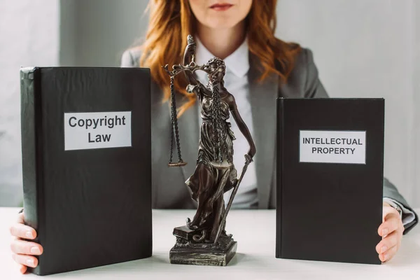 Cropped view of lawyer holding books with intellectual property and copyright law lettering near themis figurine on table on blurred background — Stock Photo