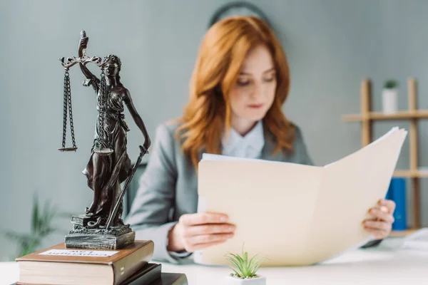 Female lawyer looking at folder, while sitting at workplace with themis figurine on pile of books on blurred background — Stock Photo