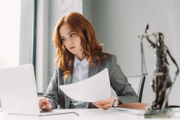 Redhead female lawyer with paper sheet looking at laptop at workplace with blurred themis figurine on foreground — Stock Photo