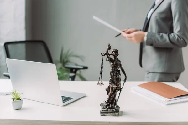 Vue recadrée de l'avocat avec feuille de papier debout près du lieu de travail avec ordinateur portable sur fond flou — Photo de stock