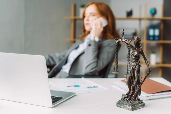 Estatuilla de Themis en la mesa cerca del ordenador portátil con abogado femenino borroso hablando por teléfono en el fondo - foto de stock