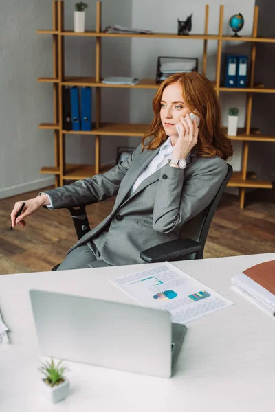 Avocate confiante parlant sur téléphone portable, tout en étant assise près du lieu de travail avec un ordinateur portable et de la paperasserie sur le premier plan flou — Photo de stock