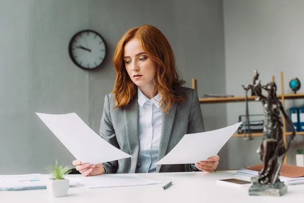 Die rothaarige Anwältin blickt auf ein Blatt Papier, während sie am Arbeitsplatz sitzt, mit verschwommener Themis-Figur im Vordergrund — Stockfoto