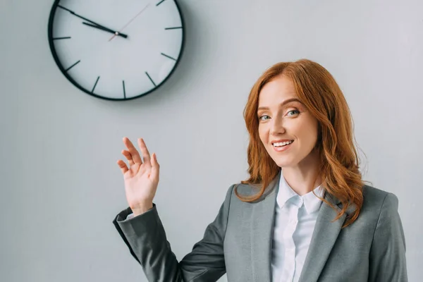 Donna d'affari sorridente in abbigliamento formale che punta con mano a orologio da parete su grigio — Foto stock