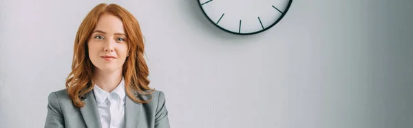 Positive redhead businesswoman looking at camera near wall clock on grey, banner — Stock Photo