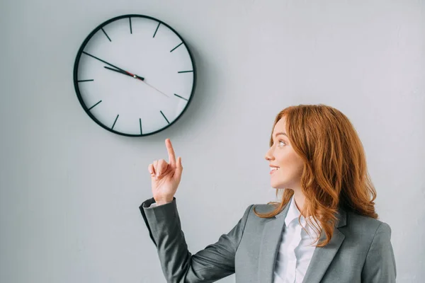 Femme d'affaires rousse souriante pointant du doigt et regardant l'horloge murale sur gris — Photo de stock