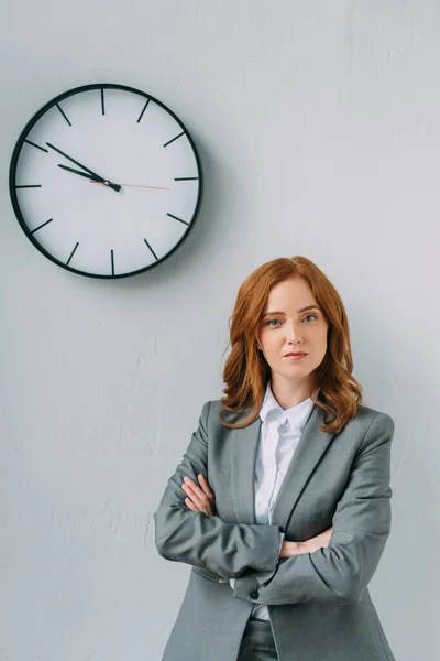 Vista frontal de empresária confiante com braços cruzados olhando para a câmera perto do relógio de parede em cinza — Fotografia de Stock