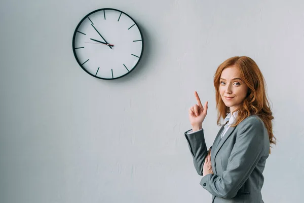 Sorridente donna d'affari rossa che punta con il dito all'orologio da parete, mentre guarda la fotocamera sul grigio — Foto stock