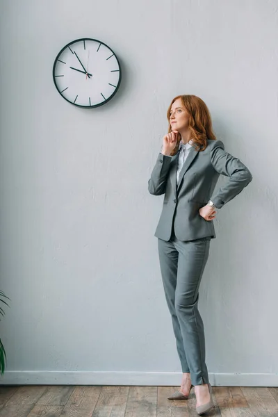 Longitud completa de la mujer de negocios reflexiva con la mano en la cadera de pie cerca de reloj de pared en la oficina — Stock Photo