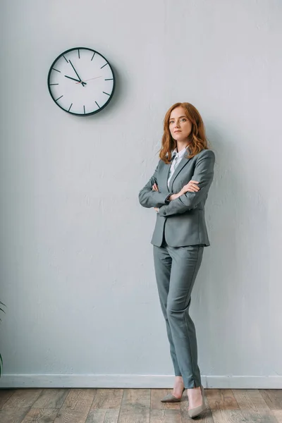 Selbstbewusste Geschäftsfrau mit verschränkten Armen steht neben Wanduhr im Büro — Stockfoto