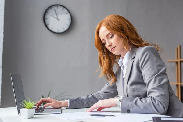 Rothaarige Geschäftsfrau schaut sich Dokumente an, während sie am Arbeitsplatz auf dem Laptop auf verschwommenem Hintergrund tippt — Stockfoto