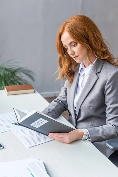 Redhead lawyer reading book with intellectual property lettering at workplace with documents on blurred background — Stock Photo