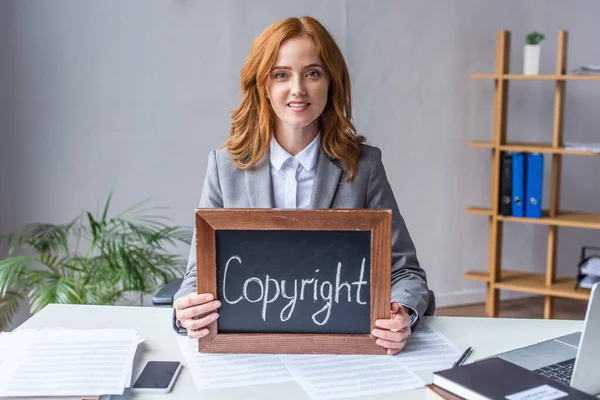 Advogado sorrindo mostrando quadro com letras de direitos autorais, enquanto sentado no local de trabalho com documentos em fundo turvo — Fotografia de Stock