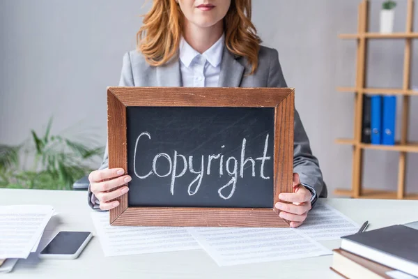 Vue recadrée de l'avocat tenant un tableau noir avec des lettres de copyright, assis à table avec des documents sur fond flou — Photo de stock