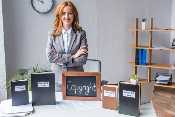 Smiling lawyer with crossed arms standing near chalkboard with copyright lettering near books on table with pile of documents — Stock Photo