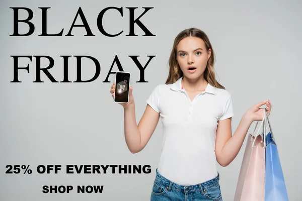 Mujer sorprendida sosteniendo teléfono inteligente y bolsas de la compra cerca del viernes negro, tienda ahora letras en gris - foto de stock