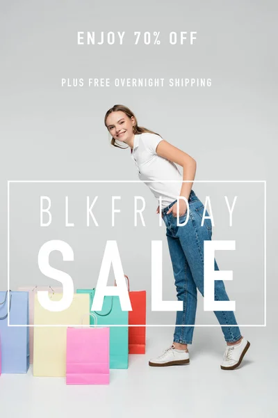 Mujer alegre de pie con las manos en los bolsillos cerca de las bolsas de la compra y blk viernes venta de letras en gris - foto de stock