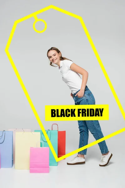 Радісна жінка стоїть з руками в кишенях біля сумки для покупок і чорна п'ятниця на сірому — стокове фото