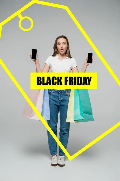 Shocked woman holding smartphones with blank screen and shopping bags near black friday lettering and yellow tag illustration on grey — Stock Photo