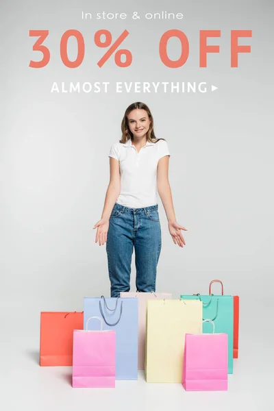 Молодая радостная женщина, стоящая и указывающая руками на сумки для покупок рядом в магазине и онлайн-надписи на сером — стоковое фото