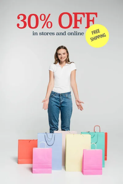 Young joyful woman standing and pointing with hands at shopping bags near free shipping lettering on grey — Stock Photo