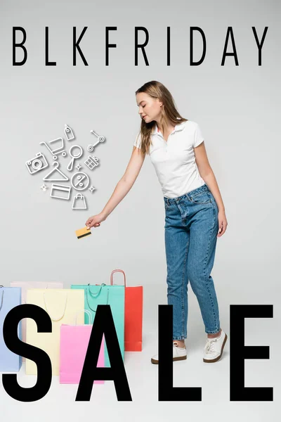Mujer joven sosteniendo la tarjeta de crédito cerca de bolsas de la compra y la ilustración y letras blk viernes en gris, concepto de viernes negro - foto de stock