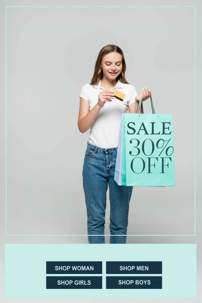 Joyful woman holding credit card and looking at blue shopping bags with sale 30 percent off lettering on grey — Stock Photo