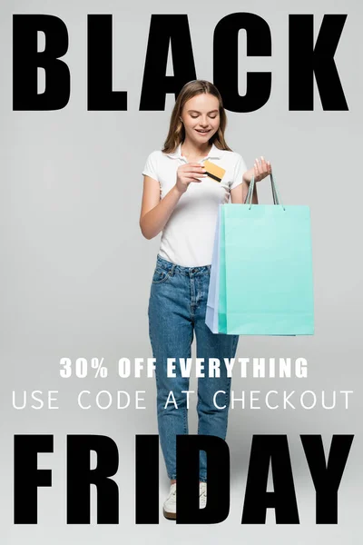 Gioiosa donna in possesso di carta di credito e guardando borse blu vicino nero venerdì lettering su grigio — Foto stock