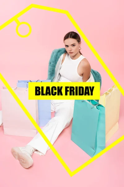 High angle view of young woman in blue faux fur jacket sitting near shopping bags and black friday lettering and tag illustration on pink — Stock Photo