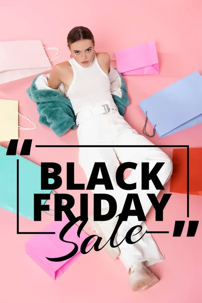 Vista aerea di giovane donna in giacca di eco-pelliccia blu seduta vicino a borse della spesa e nero venerdì vendita lettering su rosa — Foto stock