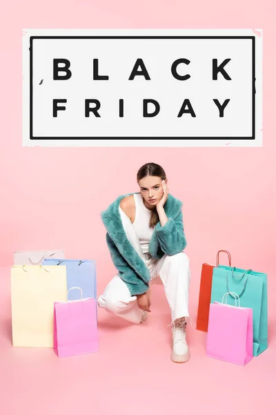 Jovem mulher em casaco de pele falso sentado perto de sacos de compras e preto sexta-feira lettering em rosa — Fotografia de Stock