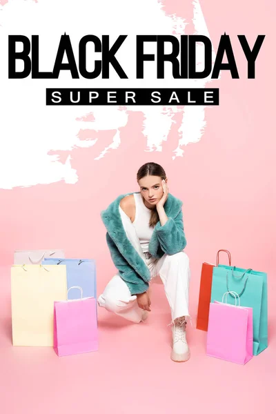 Jovem mulher em casaco de pele falso sentado perto de sacos de compras e preto sexta-feira super venda lettering em rosa — Fotografia de Stock