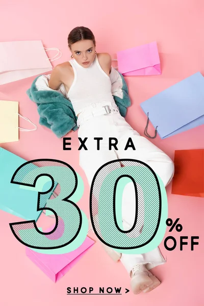 Overhead view of young woman in blue faux fur jacket sitting near shopping bags and extra 30 percent off lettering on pink — Stock Photo