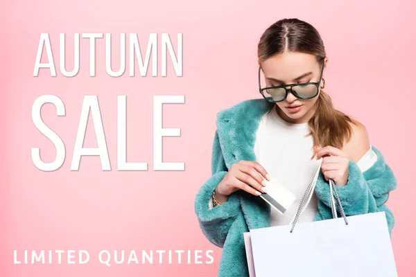 Donna alla moda in occhiali da sole in possesso di carta di credito vicino alla borsa della spesa e iscrizione di vendita autunno su rosa — Foto stock