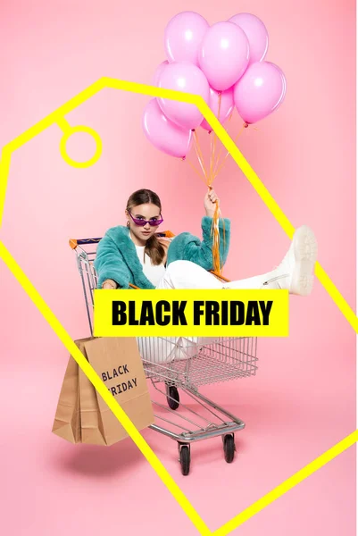 Giovane donna in occhiali da sole seduta nel carrello con le borse della spesa e tenendo palloncini vicino nero venerdì lettering e tag su rosa — Foto stock