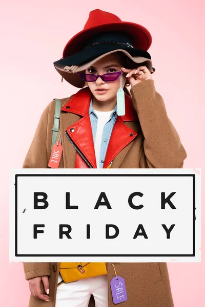 Jeune femme à la mode en chapeaux touchant lunettes de soleil avec étiquette de vente près de noir vendredi lettrage sur rose — Photo de stock
